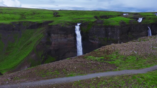 Aerial sweep over Haifoss and Granni in Thjorsardalur, Iceland, with hikers on a cliff path, basalt canyon walls, mist, and overcast light highlighting moss and strata.