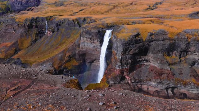 Aerial view shows Haifoss and Granni in Thjorsardalur, Iceland. Autumn colors cover the plateau, hikers stand on the rim, mist rises with a faint rainbow visible.