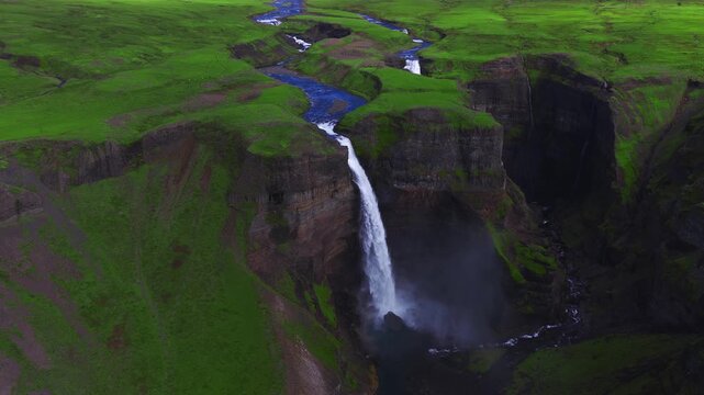 Aerial drone shot shows Haifoss and Granni in Thjorsardalur, Iceland. Mist rises as water drops into a basalt canyon, a blue river winds, summer light adds contrast. Stranger Things waterfall.