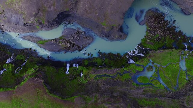 Aerial top down view shows milky turquoise meltwater braiding through dark volcanic rock and green moss in Sigoldugljufur, Iceland, with small waterfalls in daylight.