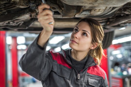 A young woman is working under a car in a garage. She is using a tool to inspect the vehicles components. The scene is bright and showcases her focused expression