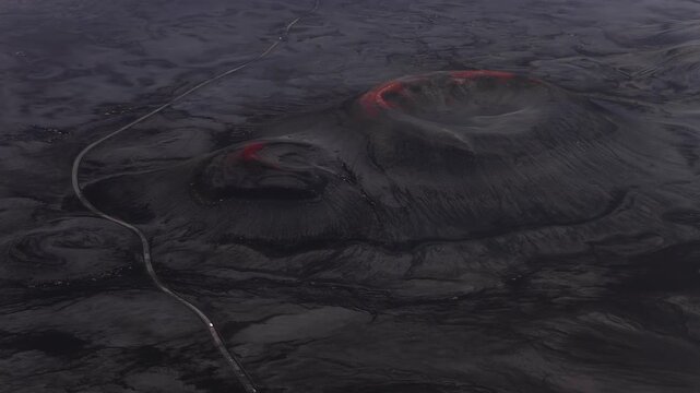 Aerial camera pans over two craters and black lava in Iceland. Ember red glows on rims, a winding road cuts through, small cars move, muted charcoal tones stand out.