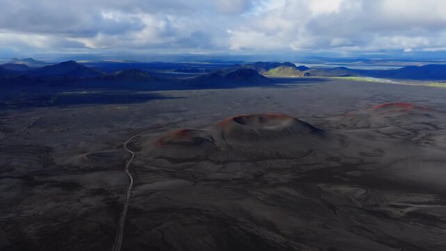 Aerial view shows red rim cinder cones on black lava and ash, a winding dirt road, and distant mountains and lakes in Iceland, under soft daylight with slow pan.