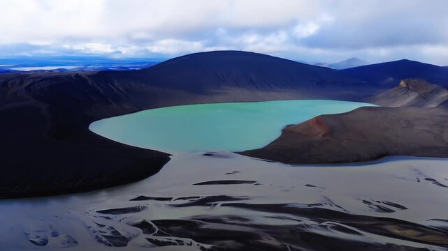 Aerial view shows a milky turquoise crater lake in Iceland in a dark caldera, black slopes, a small reddish cinder cone, and braided outwash on a black sand plain under overcast light.