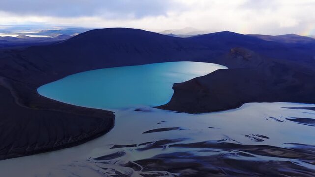 Aerial pan shows a turquoise crater lake, dark volcanic ridges, and braided glacial channels in Iceland highlands under soft overcast light with a faint rainbow.