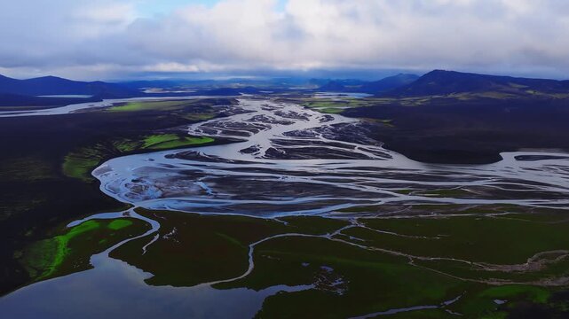 Aerial drone view shows braided glacial river, dark lava sands, green moss, sinuous channels, sandbars, and turquoise lakes in the Icelandic highlands under clouds.