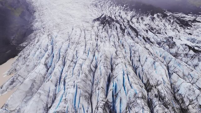 Aerial view shows an Icelandic glacier with tight tongues, deep crevasses, and a milky meltwater pool, framed by moraines and foggy highlands in soft overcast light.