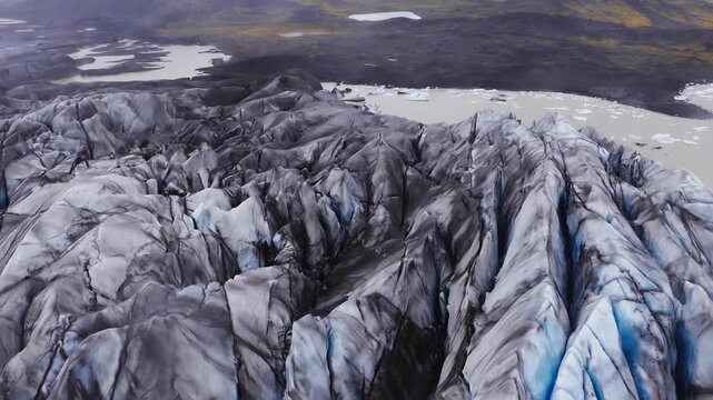Aerial view moves over a crevassed Iceland glacier with ash and blue ice, ending at a milky lagoon with drifting bergs in southeast Iceland, cool muted tones.