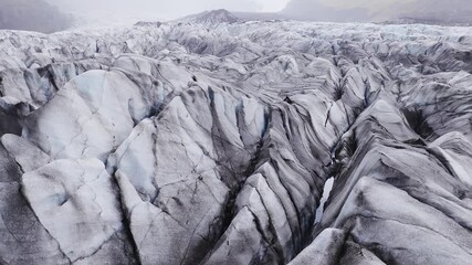 Aerial drone pan shows an Iceland glacier with jagged ridges, deep crevasses, ash and moraine streaks, turquoise meltwater holes, and overcast daylight tones. - Powered by Adobe