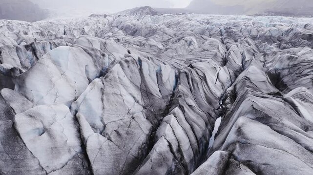 Aerial drone pan shows an Iceland glacier with jagged ridges, deep crevasses, ash and moraine streaks, turquoise meltwater holes, and overcast daylight tones.