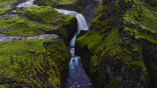 Aerial view shows a waterfall dropping into a moss clad basalt canyon in Iceland, with turquoise glacial water, mist, tiny visitors, and soft overcast light.