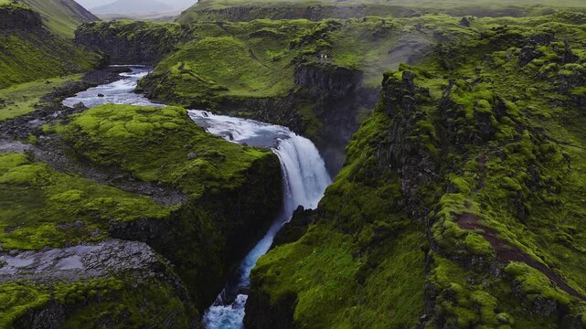 Aerial view shows a waterfall plunging into a basalt canyon in rural Iceland, with moss clad lava fields, jagged cliffs, a small viewing platform, and hikers for scale