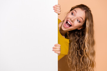 Excited young woman peeking playfully around a blank white board with a happy expression in a...