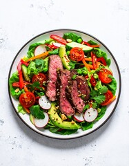 A vibrant steak salad in a white bowl. The salad contains sliced steak, vegetables like tomatoes, avocado, and lettuce, and appears to be seasoned with nuts or seeds.