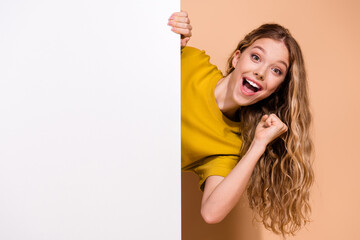 Joyful young woman in yellow shirt peeking from behind board against orange background