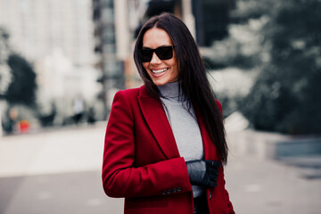 Fototapeta premium Stylish woman in a red coat and sunglass enjoying a day outdoors in an urban setting during a sunny autumn