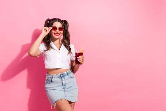 Fototapeta Playful young woman in pink studio wearing white crop top and denim skirt smiles with juice and sunglass