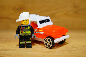 Obraz premium A Lego firefighter stands next to a toy fire truck on a wooden surface.