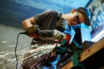 Low angle portrait of modern young mechanic working in garage, focus on sparks flying from under sending machine