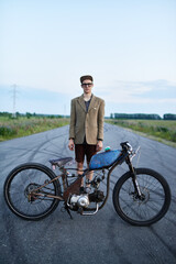 Portrait of young man wearing old-fashioned clothes standing in the middle of empty road at dusk posing neutrally with retro bike