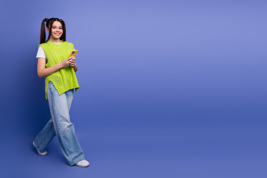 Young female student wearing a bright lime green sweater smiles while holding a smartphone against a solid blue background for lifestyle fashion and tech stock photo