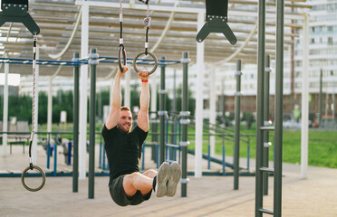 Man hanging from gym rings mid-leg lift, highlighting dynamic movement and fitness in a sunny outdoor urban park.