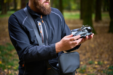 A man in a black jacket examines his drone carefully, surrounded by vibrant trees and fallen leaves in a serene park setting.