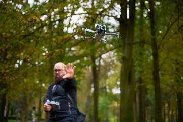 A man stands in a lush green park, his hand raised as he skillfully maneuvers a drone above him. Surrounding trees showcase vibrant autumn hues.