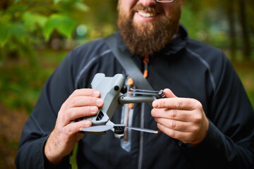 A man with a beard smiles while examining his drone, surrounded by lush trees in a peaceful forest. The warm hues of autumn enhance the tranquil atmosphere.