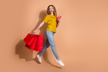 Young woman excitedly holding shopping bags and smartphone against beige background, expressing joy...