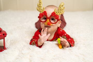 baby dressed in red Christmas pajamas wearing funny reindeer glasses with antlers and a red nose, lying on a soft white blanket next to a red lantern. Merry Christmas and a happy New Year