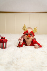 baby dressed in red Christmas pajamas wearing funny reindeer glasses with antlers and a red nose, lying on a soft white blanket next to a red lantern. Merry Christmas and a happy New Year