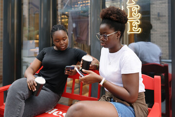 Two young women sitting outside a coffee shop with smartphones and takeaway coffee, talking and laughing, representing friendship, social connection, and modern city lifestyle.