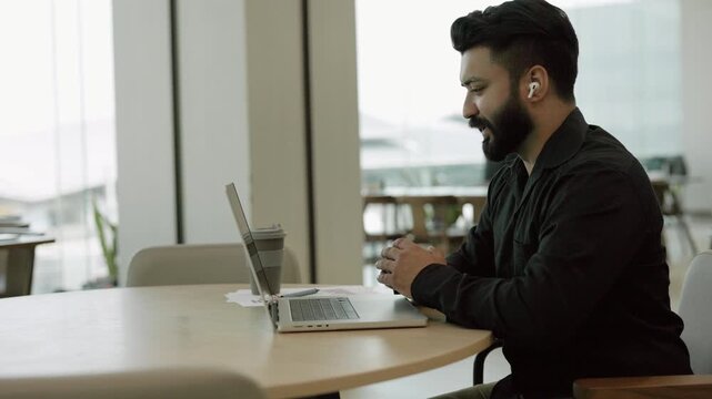 Portrait of bearded Indian man with earphones talks to colleagues at video conference in coworking. South Asian lecturer consults students at remote meeting via laptop indoors