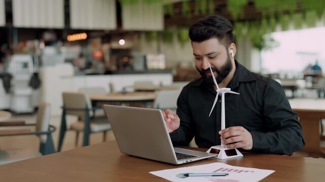 Portrait of friendly Indian technician demonstrates wind turbine model at video chat via laptop. Smiling power engineer promotes sustainable electricity at online conference
