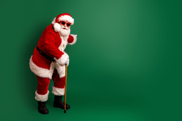 Santa in red suit with white fur hat and beard leans on a wooden staff against a green background ready for christmas shopping and holiday fun