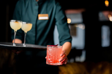 A server holding a tray with two pale cocktails while a hand in the foreground presents a pink cocktail. The scene is dimly lit and focused on the drinks and the person serving them.
