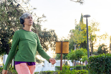 Active middle-aged woman with short gray hair and headphones walking outdoors in a park at sunset, enjoying exercise and fresh air surrounded by nature.