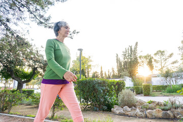 Mature woman with short gray hair and green shirt walking energetically through a park path at sunrise, surrounded by trees and morning light.