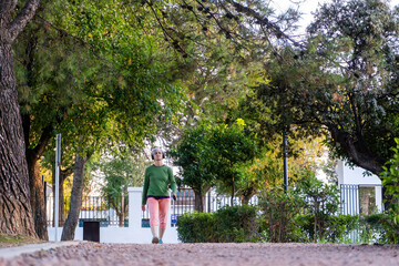 Older woman walking calmly along a shaded park path surrounded by tall trees and greenery, enjoying a peaceful outdoor moment.