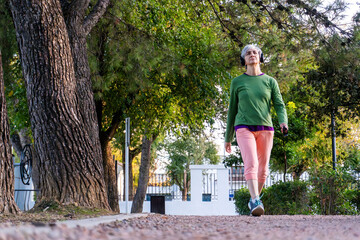 Mature woman with gray hair and sporty clothes walking through a park path lined with trees, staying active and enjoying a healthy lifestyle.