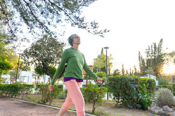 Confident woman with gray hair and headphones walking briskly along a park path surrounded by greenery, keeping an active and healthy lifestyle.