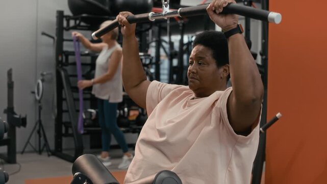 Slowmo shot of concentrated plus size senior Black woman working out in modern well-equipped gym building arm muscles using lat pulldown machine and increasing stamina for active lifestyle