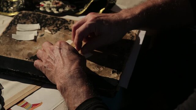 Costume designer's hands tear a paper pattern while working late, surrounded by ornate materials like a jeweled crown and embroidered belt, highlighting the craft of theatrical design.