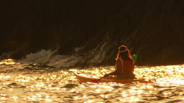 Kayaking sunset adventure woman paddles through sparkling golden water near dark cliffs enjoying summer vacation