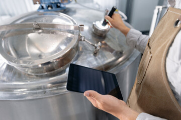 Male adult caucasian brewer operating equipment in brewery with tablet AI assistant for monitoring fresh beer
