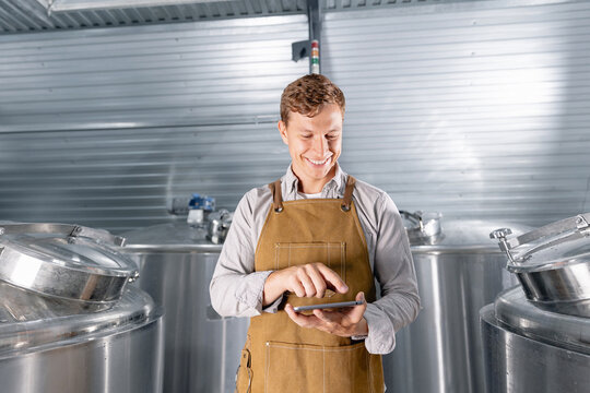 Smiling male brewer using tablet in modern brewery facility with steel tanks - Powered by Adobe