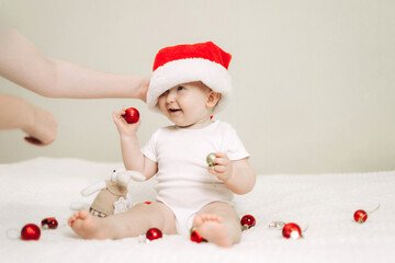 Baby wearing a Santa hat sitting on a white blanket playfully holding Christmas ornaments. An adult gently adjusts the hat, creating a festive and joyful moment.