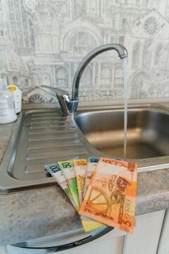 Belarusian rubles at an open water tap in the kitchen, water savings, and increased utility bills