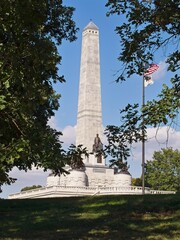Abraham Lincoln Tomb Monument in Oak Ridge Cemetery in Springfield, Illinois. Tall granite obelisk and bronze Lincoln statue under blue sky, framed by green trees and American flag. 